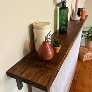 Pu&ograve; includere: Un ripiano in legno marrone scuro con un termosifone bianco dipinto dietro. Il ripiano &egrave; montato alla parete con una staffa in metallo nero. Il ripiano &egrave; decorato con un barattolo di ceramica bianca, una pera di legno, una piccola pianta grassa verde in un vaso di terracotta, una bottiglia di vetro verde con una pianta all'interno e una candela rosa.