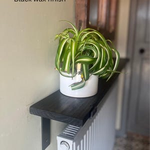 Pu&ograve; includere: Mensola in legno nero con finitura in cera nera montata su un termosifone bianco. Una pianta verde in un vaso bianco &egrave; posizionata sulla mensola. Il testo "Black wax finish" &egrave; visibile sul muro.