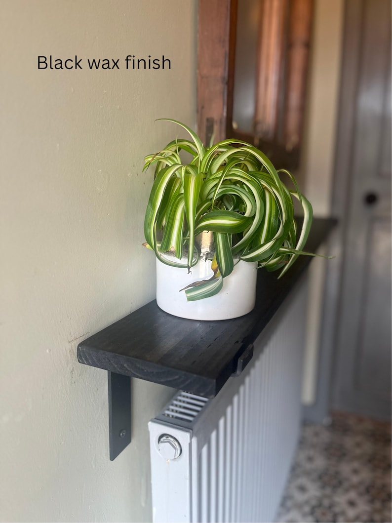 Pu&ograve; includere: Mensola in legno nero con finitura in cera nera montata su un termosifone bianco. Una pianta verde in un vaso bianco &egrave; posizionata sulla mensola. Il testo "Black wax finish" &egrave; visibile sul muro.