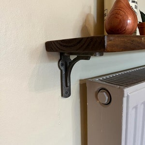 Pu&ograve; includere: Un ripiano in legno marrone scuro con una staffa in metallo nero. Il ripiano &egrave; montato su una parete bianca accanto a un radiatore bianco.