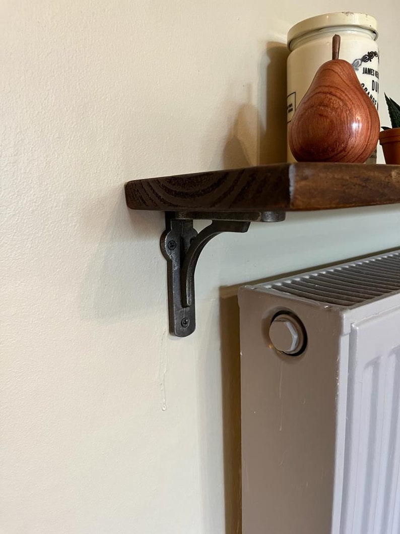 Pu&ograve; includere: Un ripiano in legno marrone scuro con una staffa in metallo nero. Il ripiano &egrave; montato su una parete bianca accanto a un radiatore bianco.