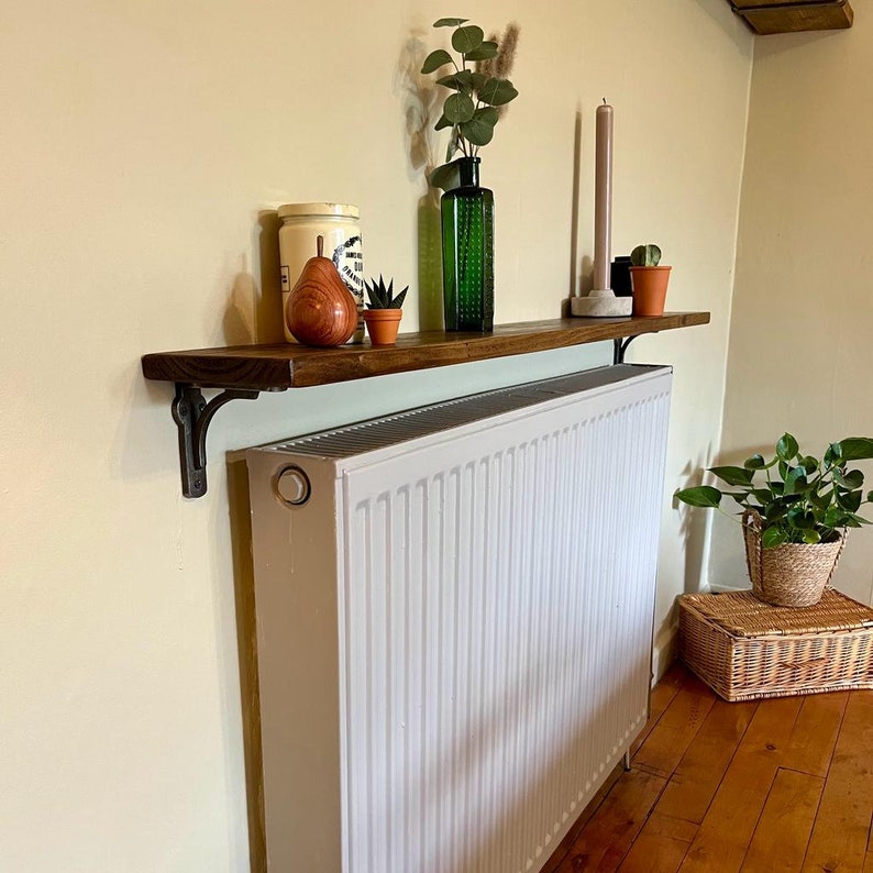 Pu&ograve; includere: Uno scaffale in legno con un termosifone bianco sotto. Lo scaffale &egrave; montato alla parete con staffe in metallo nero. Ci sono oggetti decorativi sullo scaffale, tra cui una bottiglia di vetro verde, un barattolo di ceramica, una piccola pianta in vaso e una pera di legno.