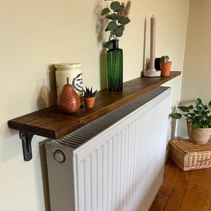 Pu&ograve; includere: Uno scaffale in legno con una tinta marrone scuro &egrave; montato sopra un radiatore bianco. Lo scaffale &egrave; decorato con un vaso di vetro verde con erbe secche, un barattolo di ceramica bianco, una piccola pianta grassa in vaso e una pera di legno.
