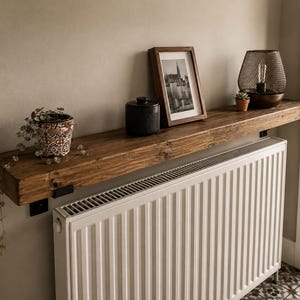 May include: A long, thick, dark-stained wooden shelf mounted above a white metal radiator. The shelf holds a patterned ceramic pot with a trailing plant, a black cylindrical container, a framed black and white photograph, and a mesh lamp.