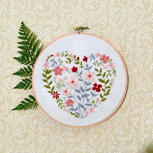 May include: A white embroidery hoop with a pink and red floral design embroidered in the shape of a heart. The hoop is on a beige background with a white floral pattern.