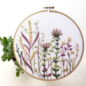 Puede incluir: Arte de bordado en aro con un diseño floral con flores y hojas rosas, amarillas y verdes. El aro es de madera y tiene un fondo de tela blanco.