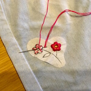 K&ouml;nnte beinhalten: Nahaufnahme eines herzf&ouml;rmigen Patches mit gestickten Blumen in rotem und schwarzem Faden. Eine Nadel mit rosa Faden ist durch den Stoff gesteckt, was auf Handn&auml;hen oder Sticken hindeutet. Der Hintergrund ist ein hellblauer Stoff.
