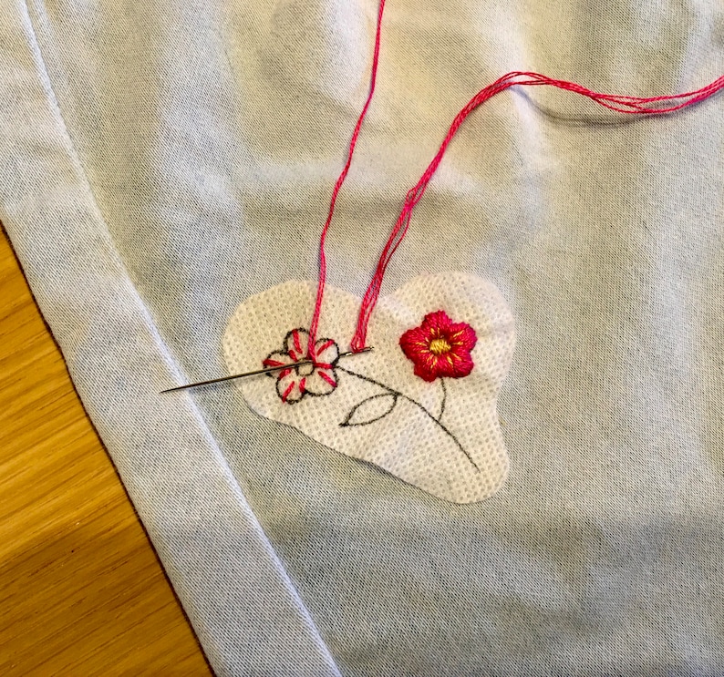 K&ouml;nnte beinhalten: Nahaufnahme eines herzf&ouml;rmigen Patches mit gestickten Blumen in rotem und schwarzem Faden. Eine Nadel mit rosa Faden ist durch den Stoff gesteckt, was auf Handn&auml;hen oder Sticken hindeutet. Der Hintergrund ist ein hellblauer Stoff.