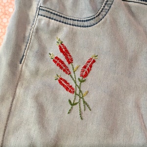 Puede incluir: Un par de pantalones vaqueros de color azul claro con un dise&ntilde;o de flores rojas bordadas en el bolsillo trasero.