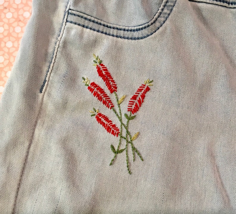 Puede incluir: Un par de pantalones vaqueros de color azul claro con un dise&ntilde;o de flores rojas bordadas en el bolsillo trasero.