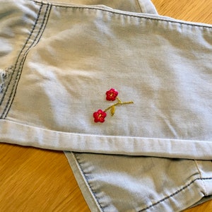 K&ouml;nnte beinhalten: Hellblaue Jeansshorts mit umgeschlagenem Saum. Die Shorts haben ein kleines gesticktes Blumendesign mit roten Bl&uuml;ten und goldenen Stielen. Der Denim hat ein verwaschenes, abgenutztes Aussehen.