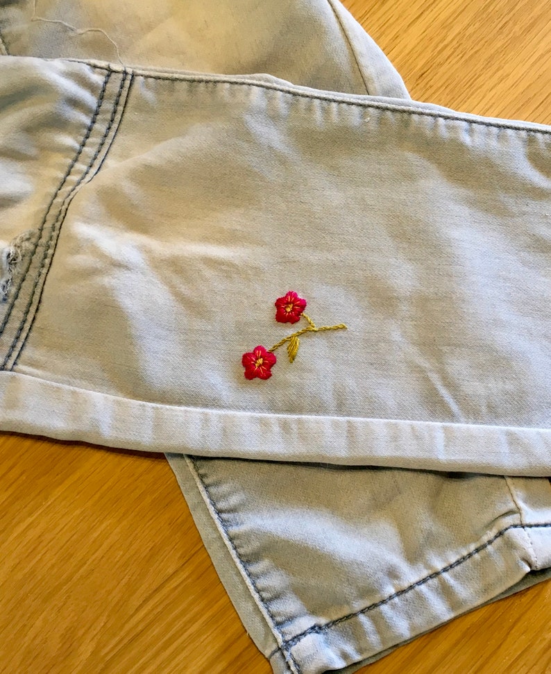 K&ouml;nnte beinhalten: Hellblaue Jeansshorts mit umgeschlagenem Saum. Die Shorts haben ein kleines gesticktes Blumendesign mit roten Bl&uuml;ten und goldenen Stielen. Der Denim hat ein verwaschenes, abgenutztes Aussehen.