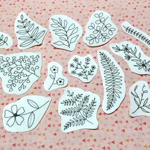 K&ouml;nnte beinhalten: Eine Sammlung wei&szlig;er Stoffflicken mit schwarzen, gestickten botanischen Designs, darunter Bl&auml;tter, Blumen und Farne. Die Flicken variieren in Gr&ouml;&szlig;e und Form und sind auf einer rosa-wei&szlig; gemusterten Oberfl&auml;che angeordnet.