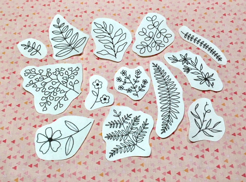 K&ouml;nnte beinhalten: Eine Sammlung wei&szlig;er Stoffflicken mit schwarzen, gestickten botanischen Designs, darunter Bl&auml;tter, Blumen und Farne. Die Flicken variieren in Gr&ouml;&szlig;e und Form und sind auf einer rosa-wei&szlig; gemusterten Oberfl&auml;che angeordnet.