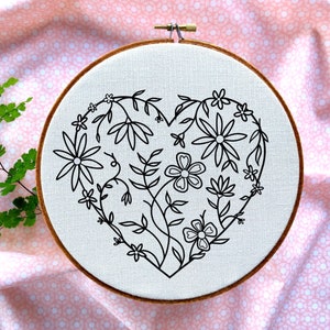 Puede incluir: Un aro de bordado con un diseño floral en forma de corazón. El aro está sobre una tela de color rosa y blanco con un patrón.