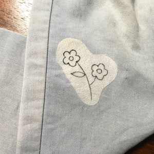 K&ouml;nnte beinhalten: Nahaufnahme von hellblauem Denim-Stoff mit einem wei&szlig;en Aufn&auml;her mit einem handgezeichneten Blumendesign. Der Aufn&auml;her hat die Form eines Herzens. Die Blumen sind schwarz umrandet.