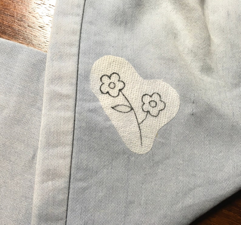 K&ouml;nnte beinhalten: Nahaufnahme von hellblauem Denim-Stoff mit einem wei&szlig;en Aufn&auml;her mit einem handgezeichneten Blumendesign. Der Aufn&auml;her hat die Form eines Herzens. Die Blumen sind schwarz umrandet.