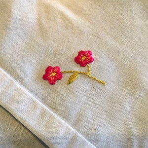 K&ouml;nnte beinhalten: Nahaufnahme eines hellblauen Stoffes mit einer detaillierten Stickerei von zwei roten Blumen mit gelben Zentren und Stielen. Das florale Design ist pr&auml;zise gestickt und verleiht dem Textil eine zarte Note.