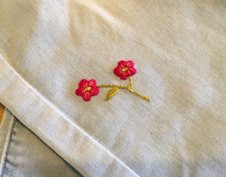 K&ouml;nnte beinhalten: Nahaufnahme eines hellblauen Stoffes mit einer detaillierten Stickerei von zwei roten Blumen mit gelben Zentren und Stielen. Das florale Design ist pr&auml;zise gestickt und verleiht dem Textil eine zarte Note.
