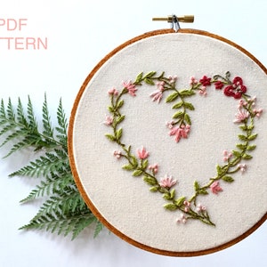 Puede incluir: Diseño de corazón floral bordado en un aro. El diseño presenta flores y hojas rosas y verdes, con algunas flores rojas en el lado derecho.
