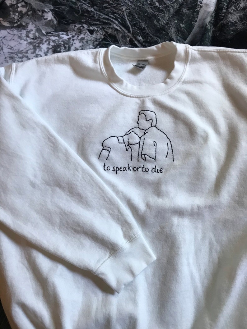 Puede incluir: Sudadera blanca con un dise&ntilde;o bordado en negro de dos personas mirando hacia arriba y el texto "to speak or to die".