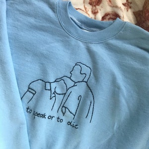 Puede incluir: Sudadera azul claro con un dise&ntilde;o bordado en negro de tres personas y el texto "to speak or to die".