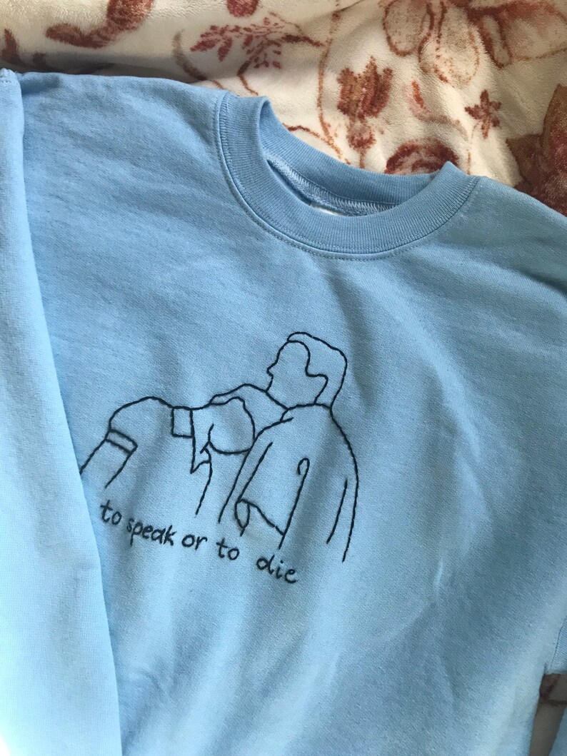 Puede incluir: Sudadera azul claro con un dise&ntilde;o bordado en negro de tres personas y el texto "to speak or to die".