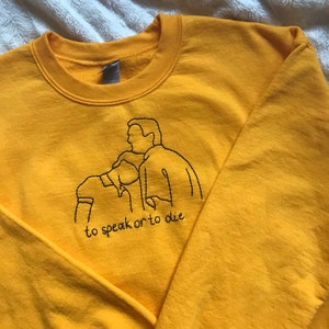 Puede incluir: Sudadera amarilla con un dise&ntilde;o bordado en negro de dos personas y el texto "to speak or to die".