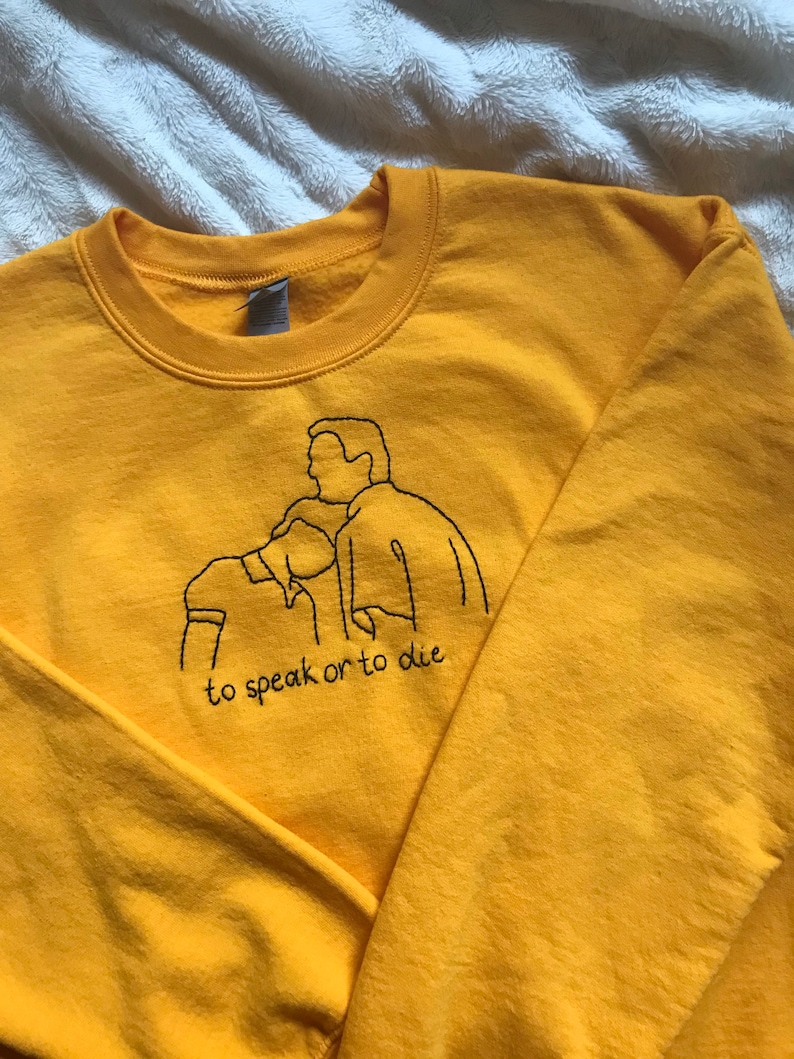 Puede incluir: Sudadera amarilla con un dise&ntilde;o bordado en negro de dos personas y el texto "to speak or to die".