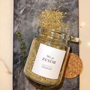 May include: A glass jar of Za'atar seasoning with a white label, sitting on a dark gray marble cutting board. The label reads "No. 40 Za'atar Seasoning". A small sprig of thyme and a cork are also present.