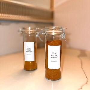 May include: Two clear glass spice jars with metal clasps, filled with curry powder and garam masala. Each jar has a white label with black text. The jars are on a white countertop.