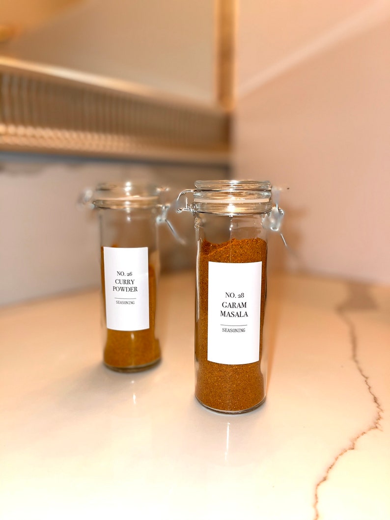 May include: Two clear glass spice jars with metal clasps, filled with curry powder and garam masala. Each jar has a white label with black text. The jars are on a white countertop.