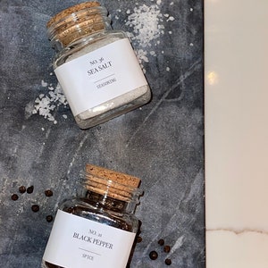 May include: Two small glass jars with cork lids, labeled "No. 36 Sea Salt" and "No. 21 Black Pepper." The sea salt jar is filled with white crystals, and the black pepper jar contains black peppercorns. The jars sit on a dark gray surface.