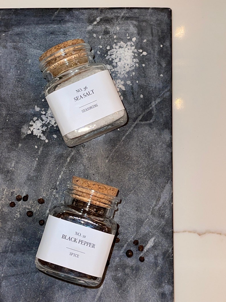 May include: Two small glass jars with cork lids, labeled "No. 36 Sea Salt" and "No. 21 Black Pepper." The sea salt jar is filled with white crystals, and the black pepper jar contains black peppercorns. The jars sit on a dark gray surface.