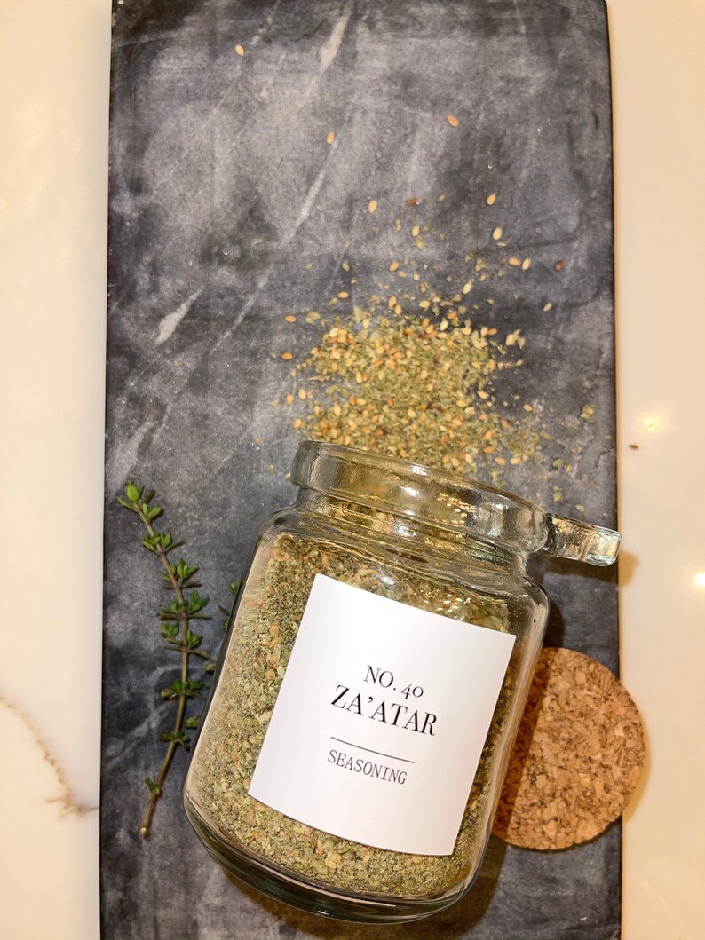 May include: A glass jar of Za'atar seasoning with a white label, sitting on a dark gray marble cutting board. The label reads "No. 40 Za'atar Seasoning". A small sprig of thyme and a cork are also present.