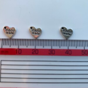 May include: Three silver heart-shaped charms with the words "hand made" inscribed on them. The charms are approximately 1 cm in size and are shown against a white background with a red ruler.