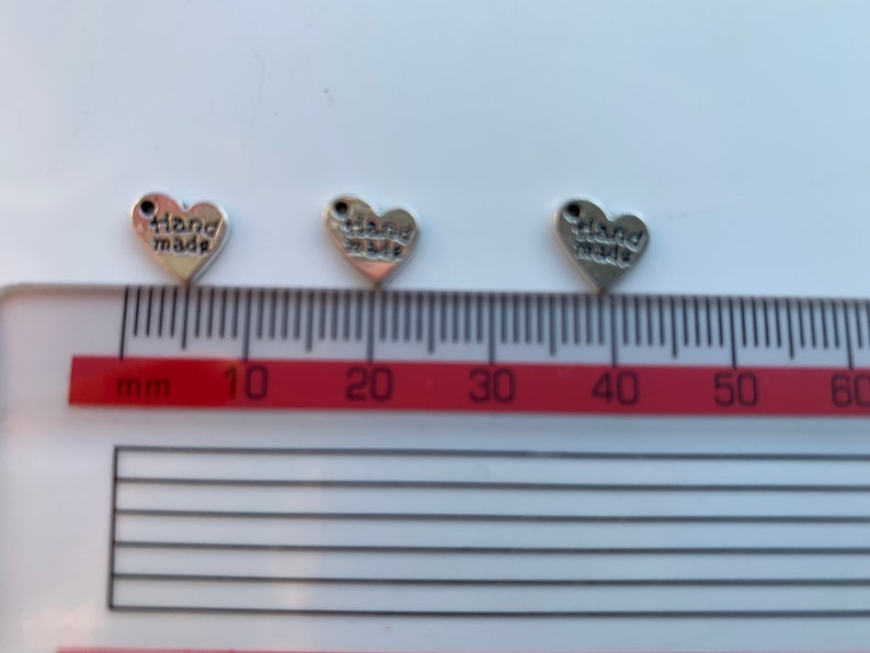 May include: Three silver heart-shaped charms with the words "hand made" inscribed on them. The charms are approximately 1 cm in size and are shown against a white background with a red ruler.