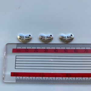 May include: Three silver heart-shaped charms with the text "Hand Made With Love" on each charm. The charms are measured on a ruler with centimeter markings.