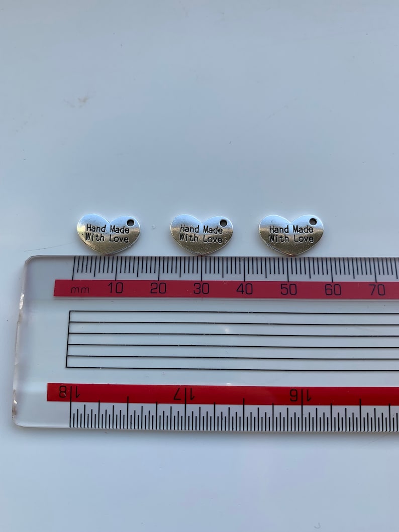 May include: Three silver heart-shaped charms with the text "Hand Made With Love" on each charm. The charms are measured on a ruler with centimeter markings.