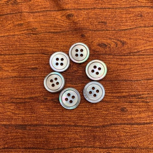 May include: Six iridescent, mother-of-pearl buttons with four holes each, arranged in a circle on a brown fabric background.