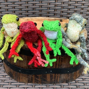 May include: Four knitted frog figurines in different colours, including green, red, and grey, sitting on a wooden barrel.