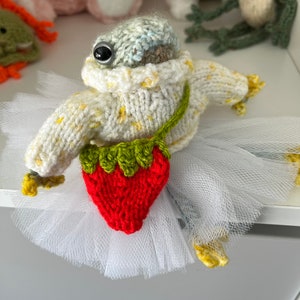 May include: A knitted frog wearing a white sweater with yellow polka dots and a red and green knitted strawberry. The frog is wearing a white tulle tutu and has a black button eye.