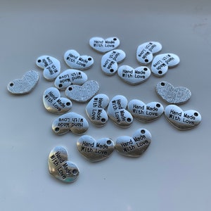 May include: A collection of silver metal charms with the text "Hand Made With Love" on each charm. The charms are in the shape of a heart and a bean.