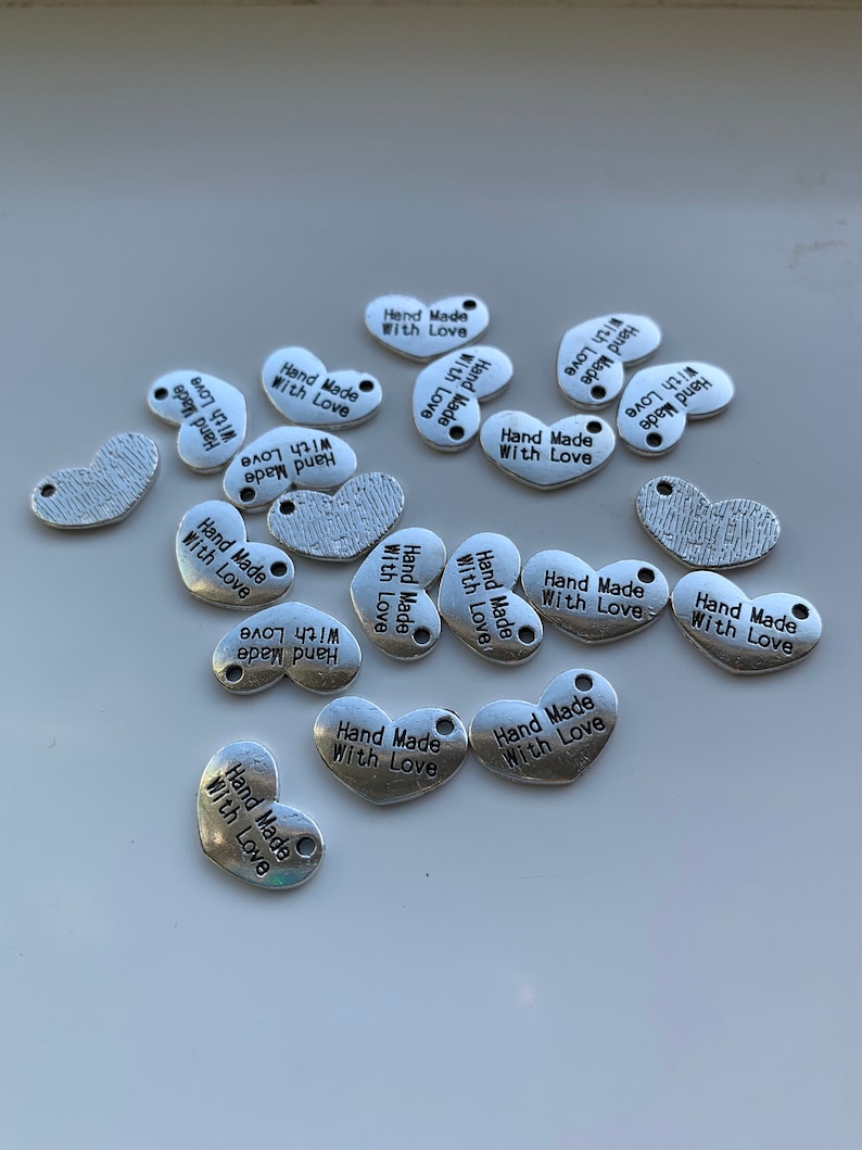May include: A collection of silver metal charms with the text "Hand Made With Love" on each charm. The charms are in the shape of a heart and a bean.