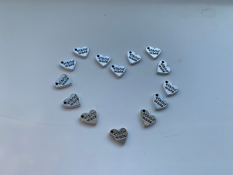 May include: A collection of silver heart-shaped charms with the words "Hand Made" printed on them.