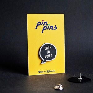 May include: Black and white enamel pin with the text "Born to Build" in a speech bubble shape. The pin is on a yellow card with the text "pin pins" and "Made in Lithuania".