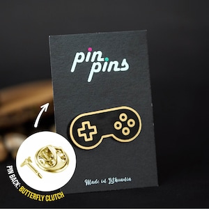 Puede incluir: Pin de color dorado con un mando de videojuego en blanco y negro. El pin tiene un cierre de mariposa en la parte posterior. El pin se muestra en una tarjeta negra con el texto "pin pins" y "Hecho en Lituania".