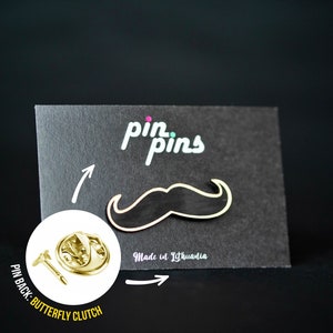 Puede incluir: Pin de esmalte negro y blanco con forma de bigote. El pin está en una tarjeta negra con el texto "pin.pins" y "Made in Lithuania". La parte posterior del pin tiene un cierre de mariposa.