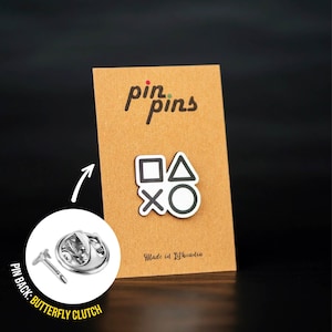 May include: A white enamel pin with black outlines of the PlayStation controller symbols: square, triangle, circle, and X. The pin is on a brown card with the text "pin pins" and "Made in Lithuania".