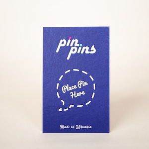 May include: A blue card with the text "pin.pins" in white with a pink and blue dot above the "i" in "pin". The card has a white dashed circle with the text "Place Pin Here" inside. The bottom of the card says "Made in Lithuania" in white.